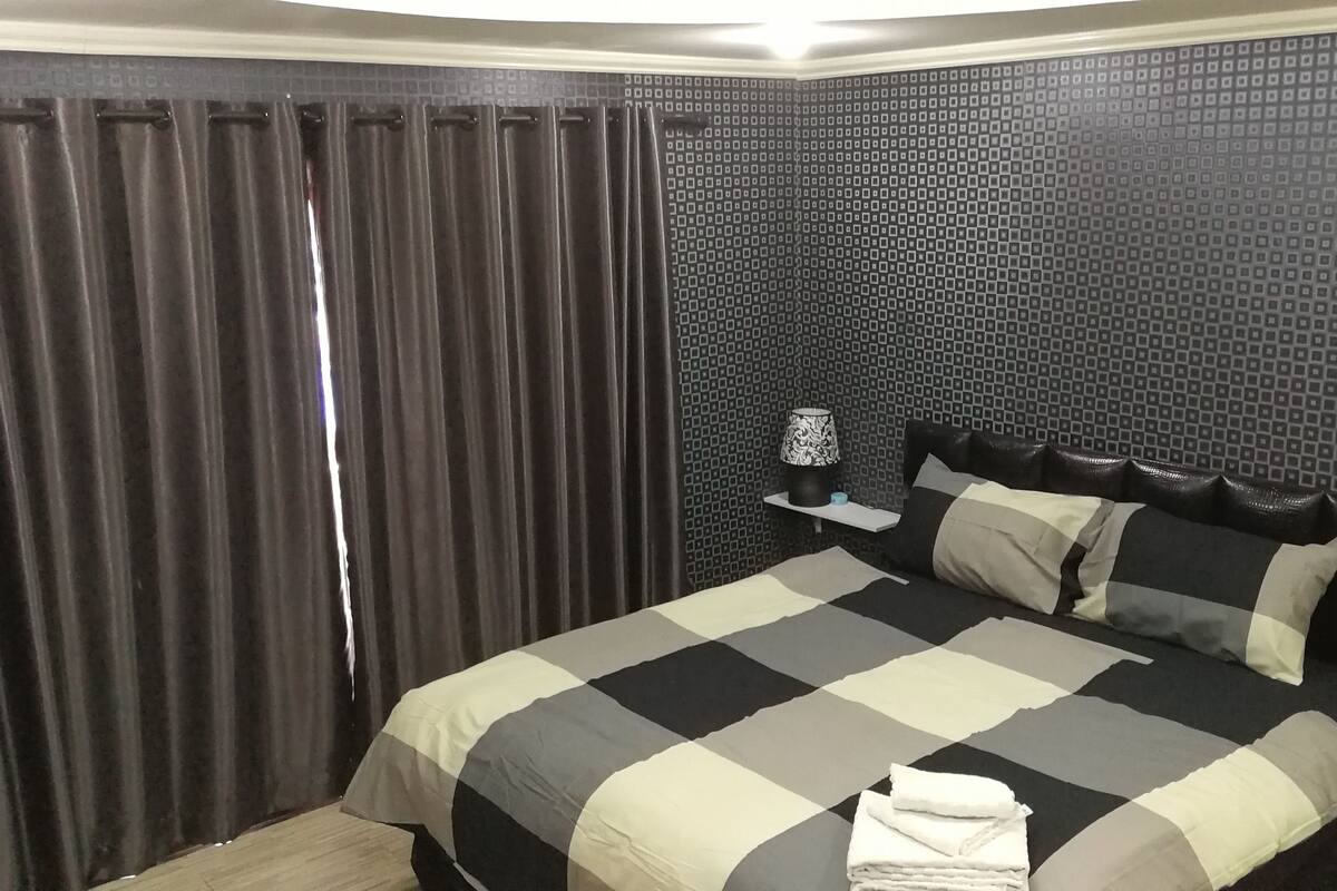 A hotel room with a bed, curtains, a patterned wall, a nightstand, and a lamp.