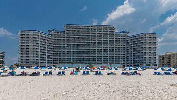 A large hotel with multiple floors and balconies, a sandy beach in the foreground with several umbrellas and lounge chairs, and a clear blue sky.