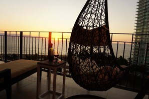 A balcony with a hanging chair, a table with a bottle and glass, and a view of the sunset over the sea.