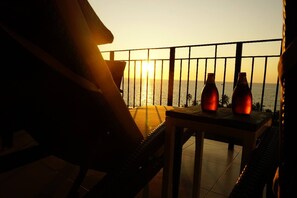 A balcony with a view of the sunset over the ocean, two bottles on a table, and a chair.