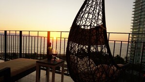 Un balcón con una silla colgante, una mesita con una botella y un vaso, y vista al atardecer sobre el agua.