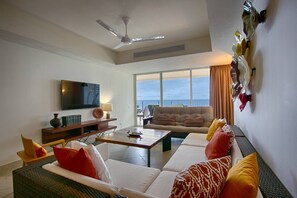 A modern living room with a flat-screen TV, a sofa set, a coffee table, and a view of the ocean through large windows.