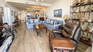 A spacious living room with a blue sofa, a leather armchair, and a coffee table. There is a ceiling fan, a bookshelf with model ships, and a painting of a lighthouse.