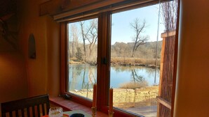 Un comedor con una mesa dispuesta para dos personas, una ventana con vistas a un lago y árboles, y una vela sobre la mesa.
