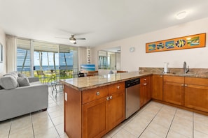 A modern kitchen with a large island, wooden cabinets, and a view of the ocean through the sliding glass doors. - Second Floor Oceanview with Gorgeous Sunsets (Kihei)