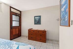 A bedroom with a wooden door, a dresser, a framed picture on the wall, and a bed with a blue and white patterned bedspread. - Second Floor Oceanview with Gorgeous Sunsets (Kihei)