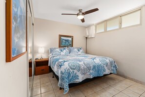 Ein Schlafzimmer mit einem Bett, einem Nachttisch, einer Lampe, einem Deckenventilator, einem Fenster mit Vorhängen und einem gerahmten Bild an der Wand.