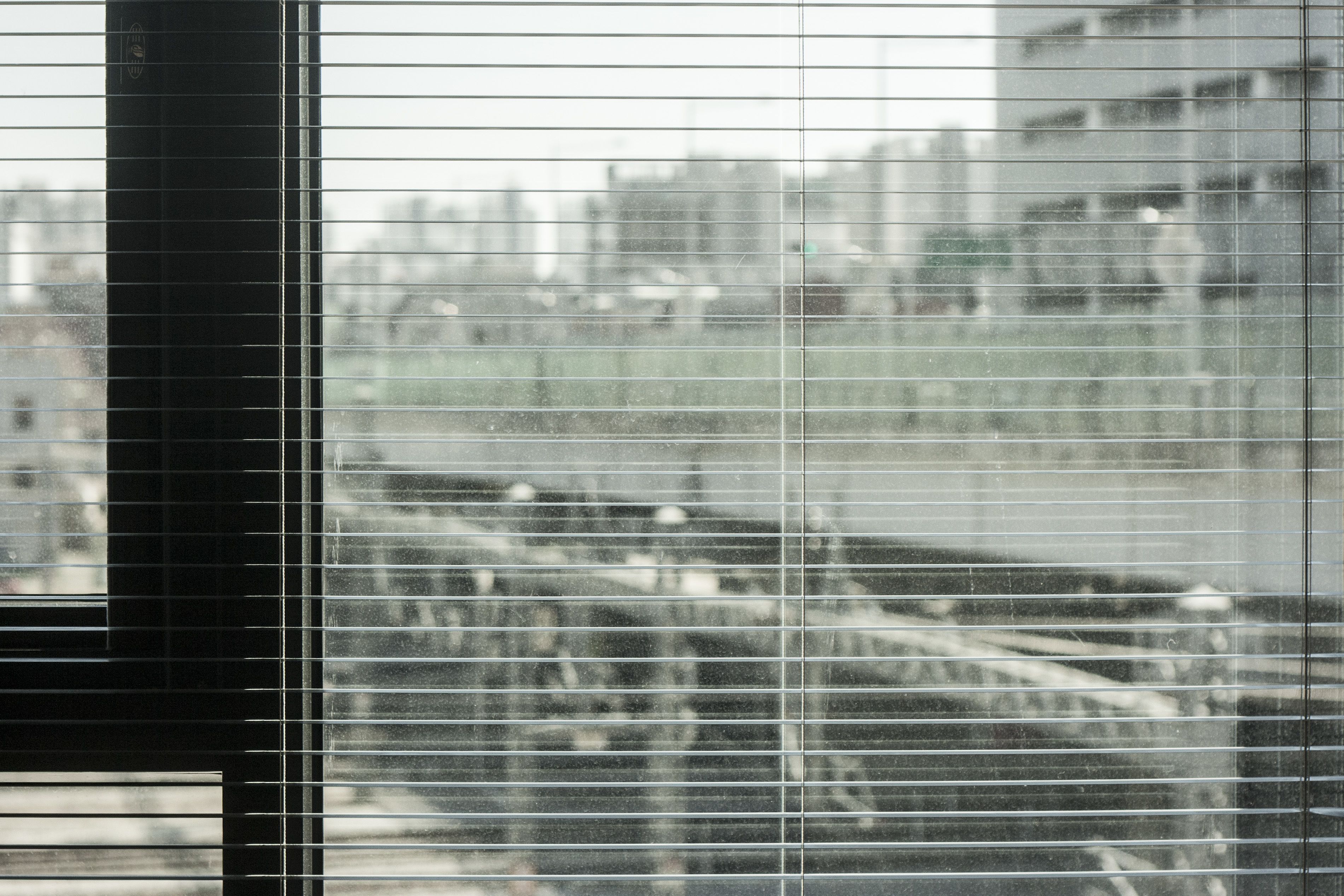 A view through a window with horizontal blinds, featuring a blurred urban landscape with buildings and a bridge.