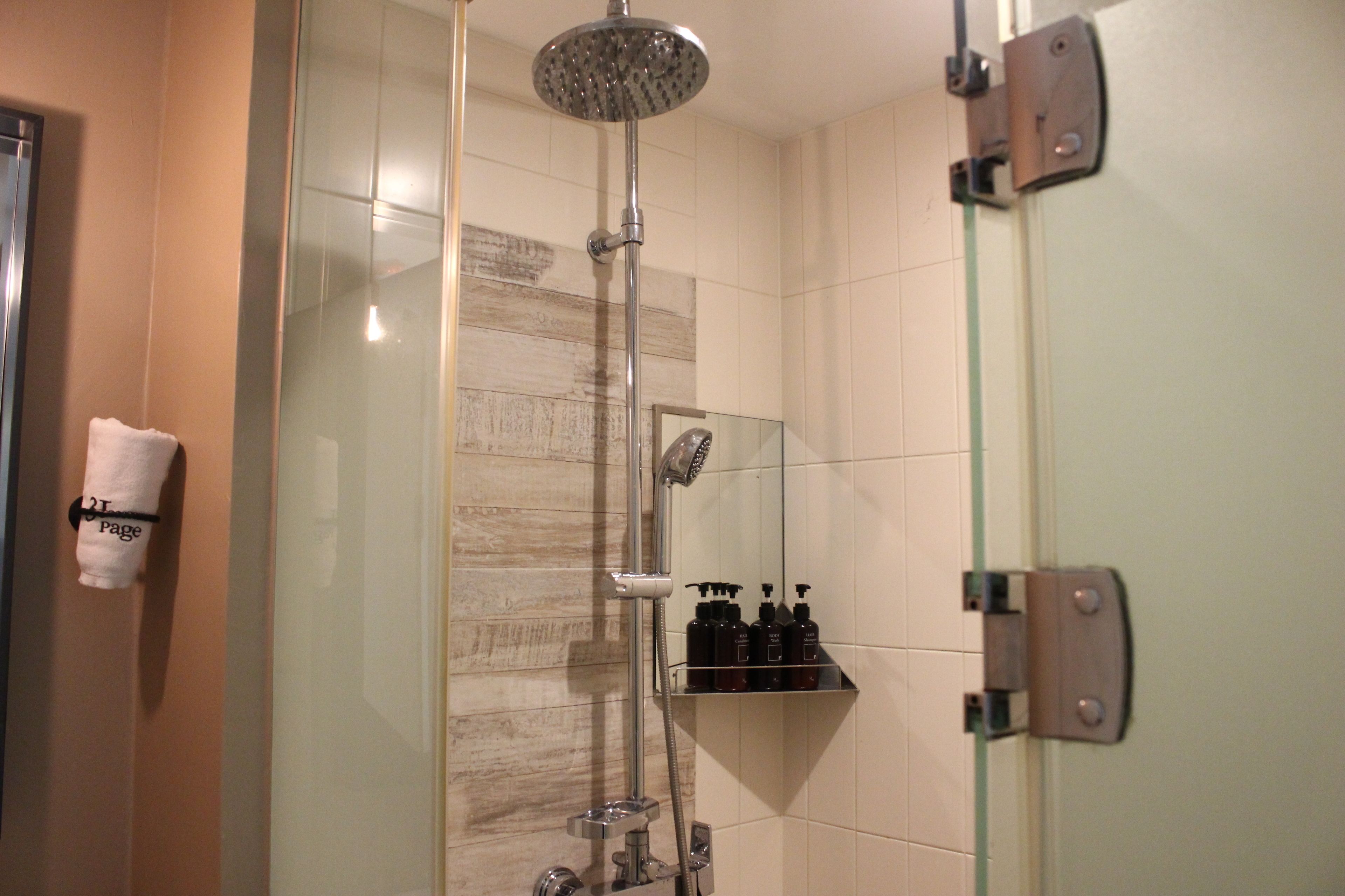 A glass-enclosed shower with a handheld showerhead, a wall-mounted faucet, and a shelf with toiletries.