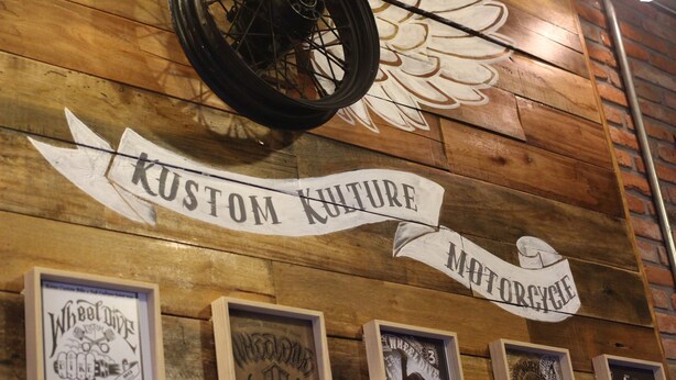 Un mur à la texture bois, une enseigne indiquant « Kustom Culture », et des œuvres d’art encadrées.