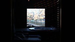 Une pièce faiblement éclairée, d’où l’on aperçoit un paysage urbain à travers une fenêtre, avec un pont et des immeubles de grande hauteur.