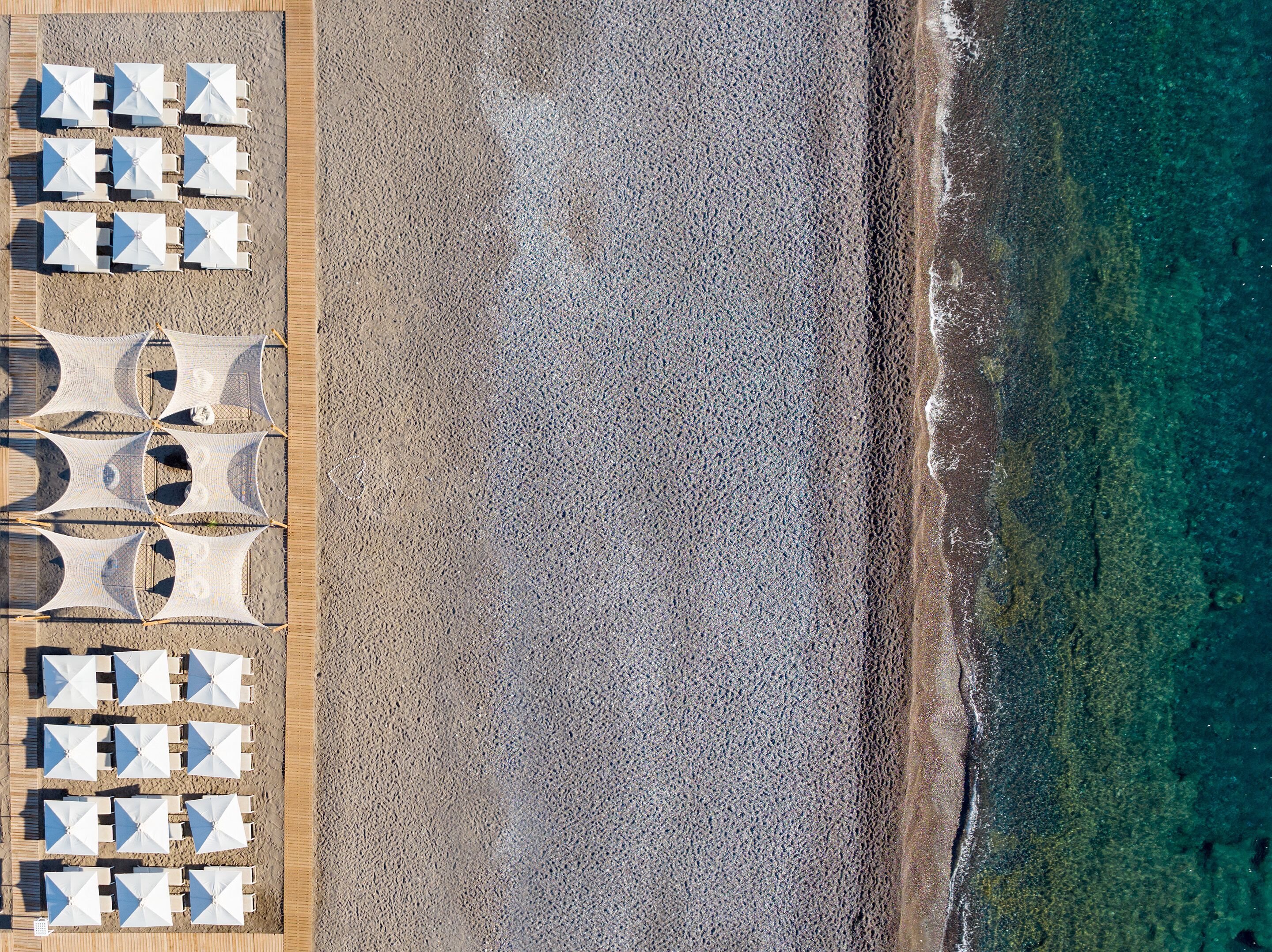 Ein Luftbild zeigt einen Strand mit Reihen aus weißen Sonnenschirmen und Liegestühlen, einer Holzpromenade und freiem Blick auf das Meer.