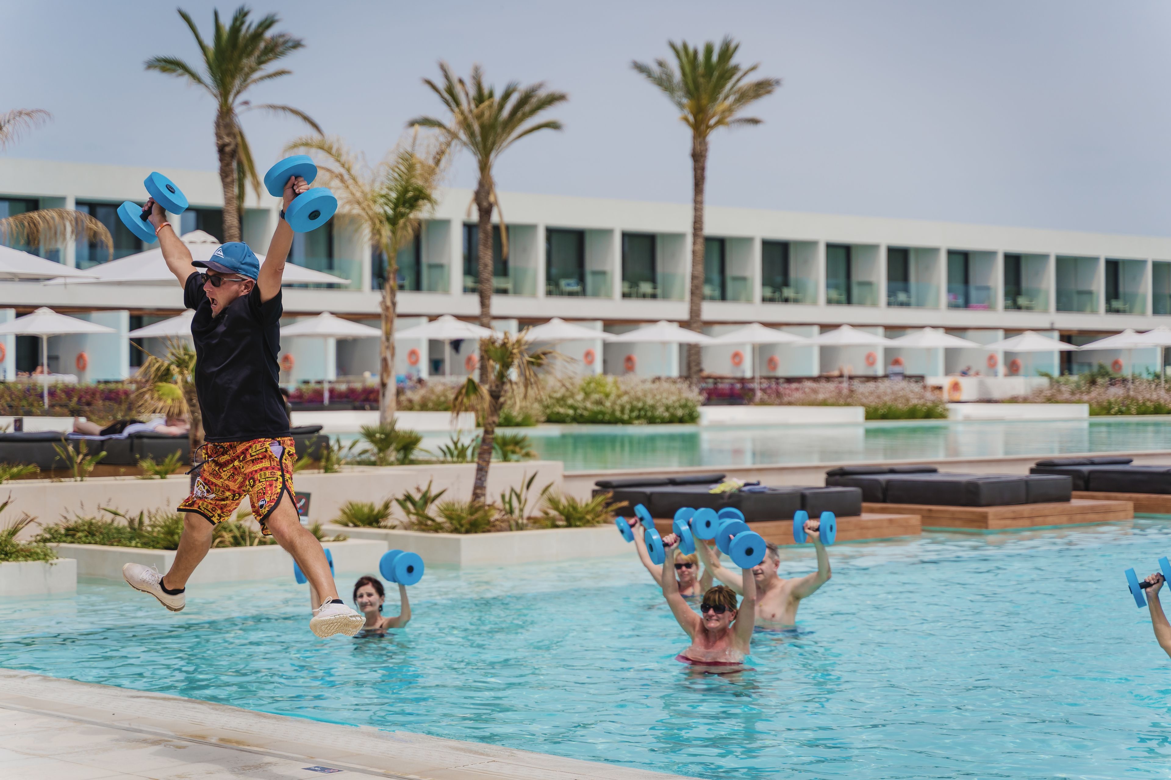 A swimming pool area with people using blue dumbbells, palm trees, and a modern building with large windows in the background.