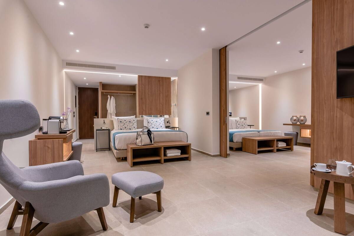 Ein modernes Hotelzimmer mit einem großen Bett, einer Sitzecke mit Sessel und Hocker, einem kleinen Tisch mit Tasse und Glas sowie einem an der Wand befestigten Fernseher.