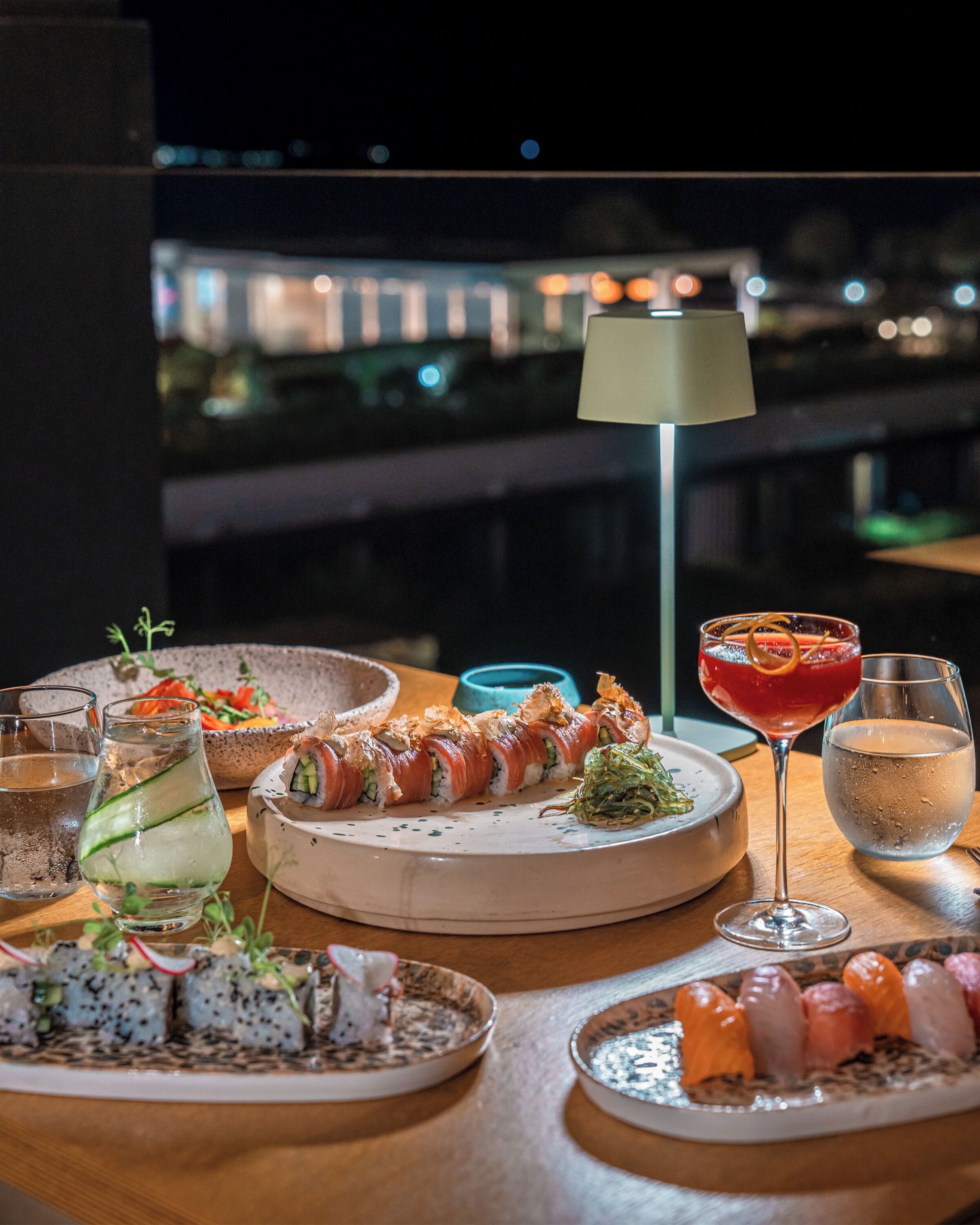 Een gedekte tafel met diverse gerechten, waaronder sushi en een cocktail, met een stadszicht op de achtergrond.