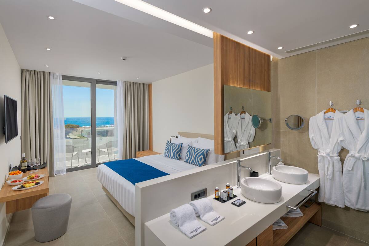 Ein modernes Hotelzimmer mit einem großen Bett, einem Badezimmer mit zwei Waschbecken, einem Spiegel und Meerblick vom Balkon.