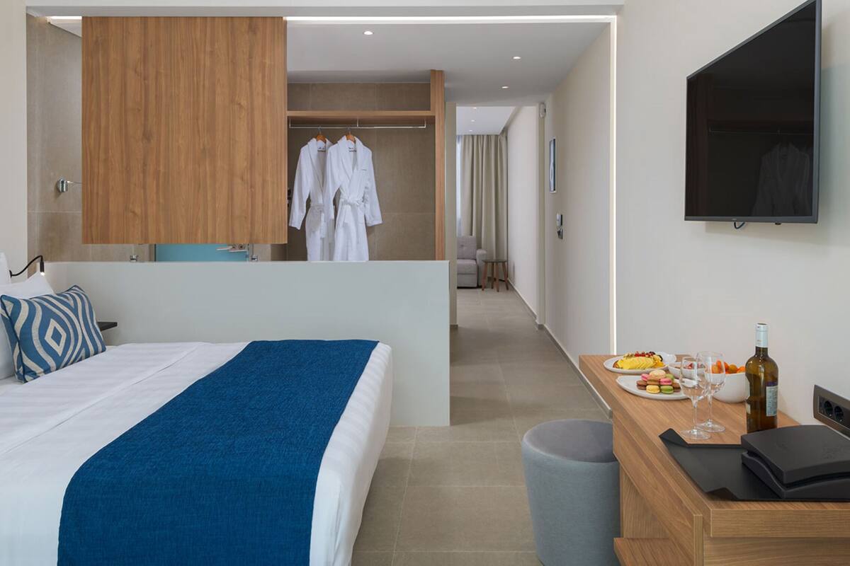 Ein Hotelzimmer mit einem Bett, einem Holzschrank, einem Spiegel, einem Fernseher, einem Schreibtisch mit einer Weinflasche und einem Papierkorb.
