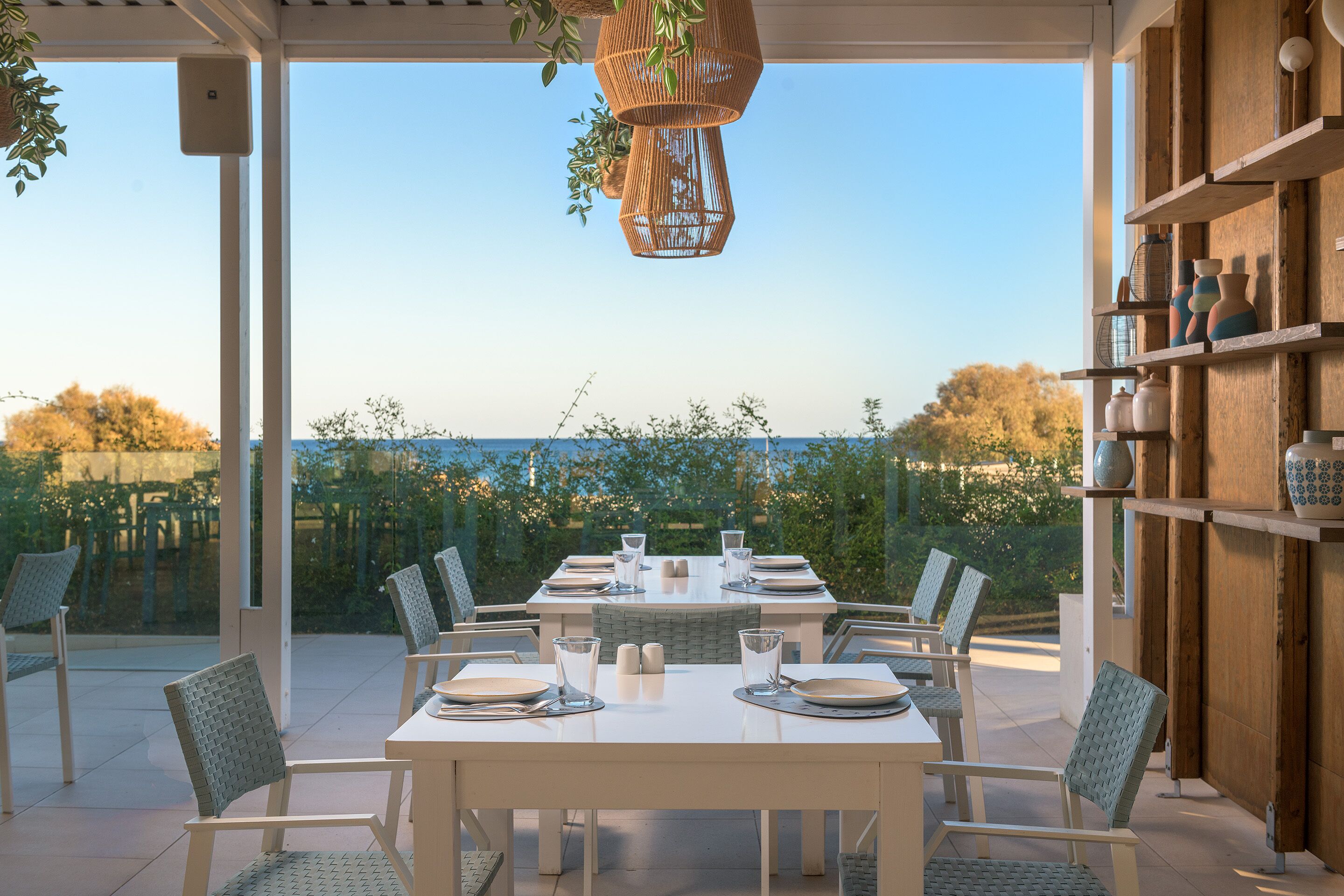 Un área de comedor al aire libre con una mesa blanca preparada para seis personas, rodeada de sillas de mimbre, con vista al mar y follaje en el fondo.