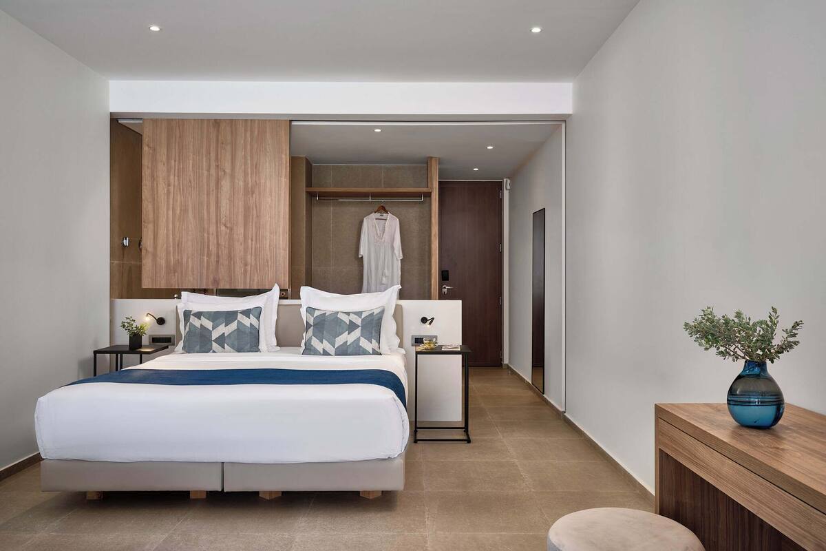 Ein modernes Hotelzimmer mit einem großen Bett, Nachttischen, einem Kleiderschrank und einem kleinen Schreibtisch mit einer Vase.