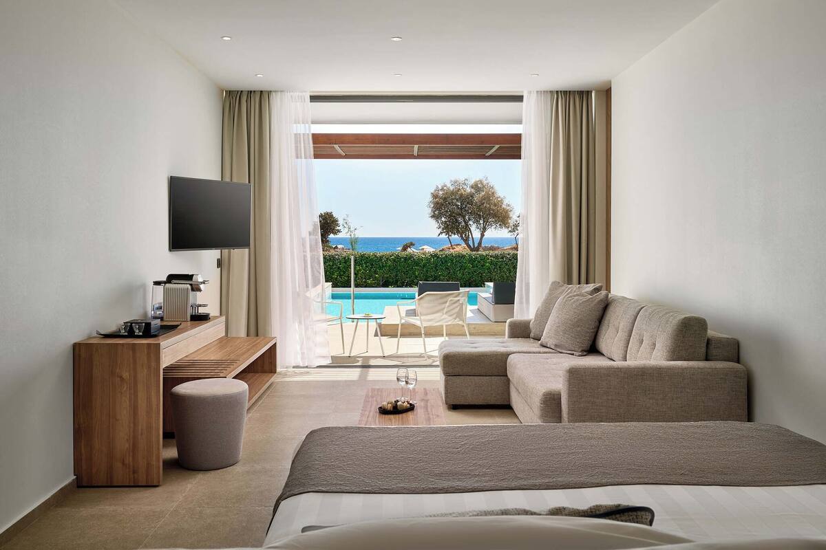 A modern hotel room with a large bed, a sofa, a TV, and a desk. There is a pool visible through the window.