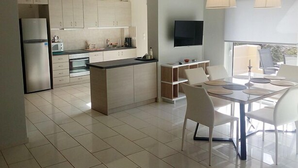 A modern kitchen with white cabinets, a central island, and a dining area with a table and chairs. - Geo Maisonette Lagonissi (Saronikos)