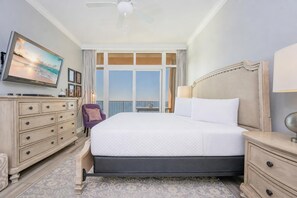 A hotel room with a large bed, a dresser, a chair, a TV, and a view of the ocean.