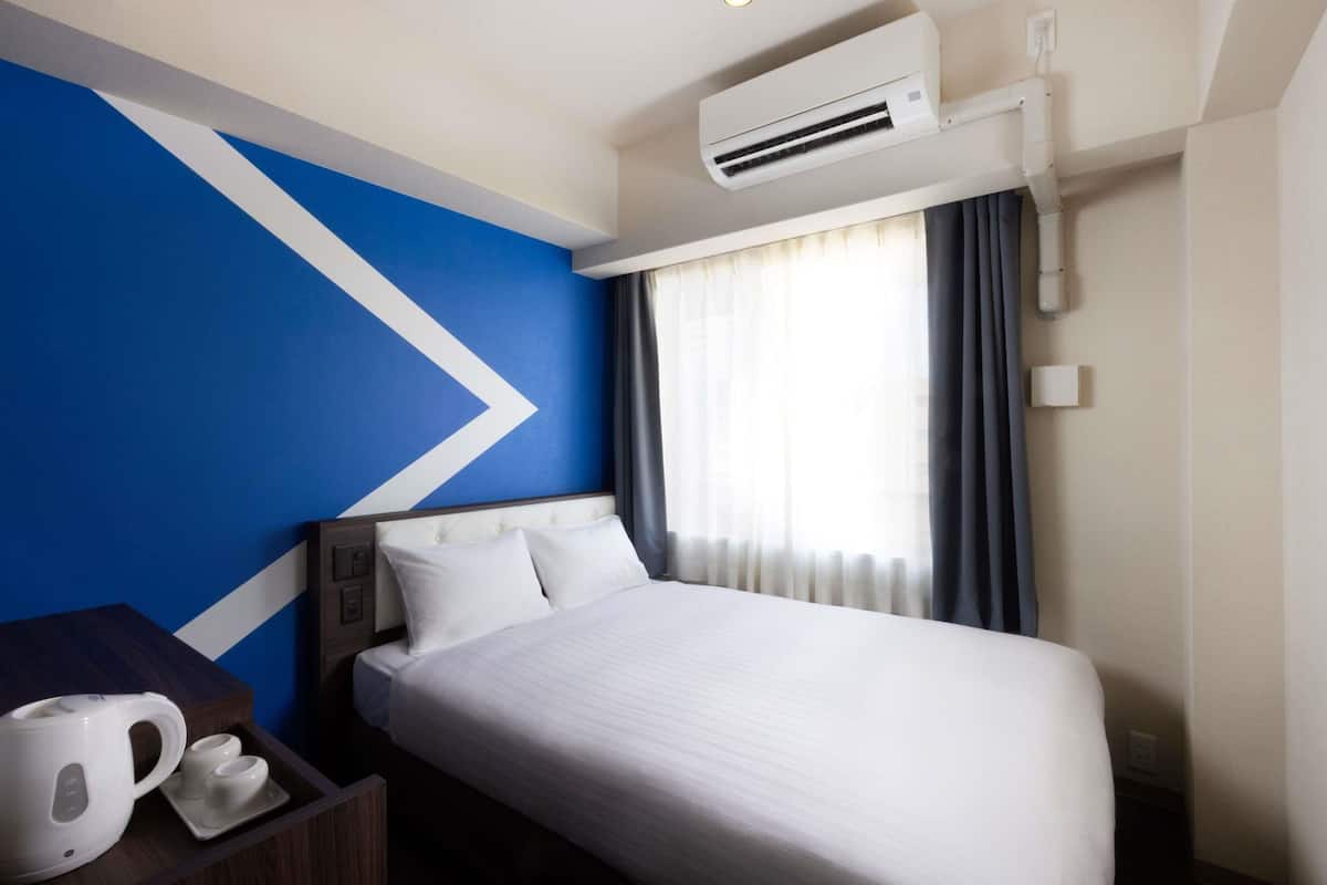 Habitación de hotel con cama, mesita de noche con tetera, aire acondicionado instalado en la pared y ventana con cortinas.