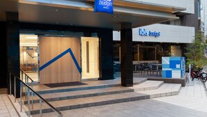 Exterior de un hotel ibis budget con un diseño moderno, destacando un prominente logo azul, una escalera que conduce a la entrada y un área de descanso visible a través de las puertas de cristal.