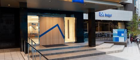 The exterior of an ibis budget hotel with a modern design, featuring a prominent blue logo, a staircase leading to the entrance, and a seating area visible through the glass doors.