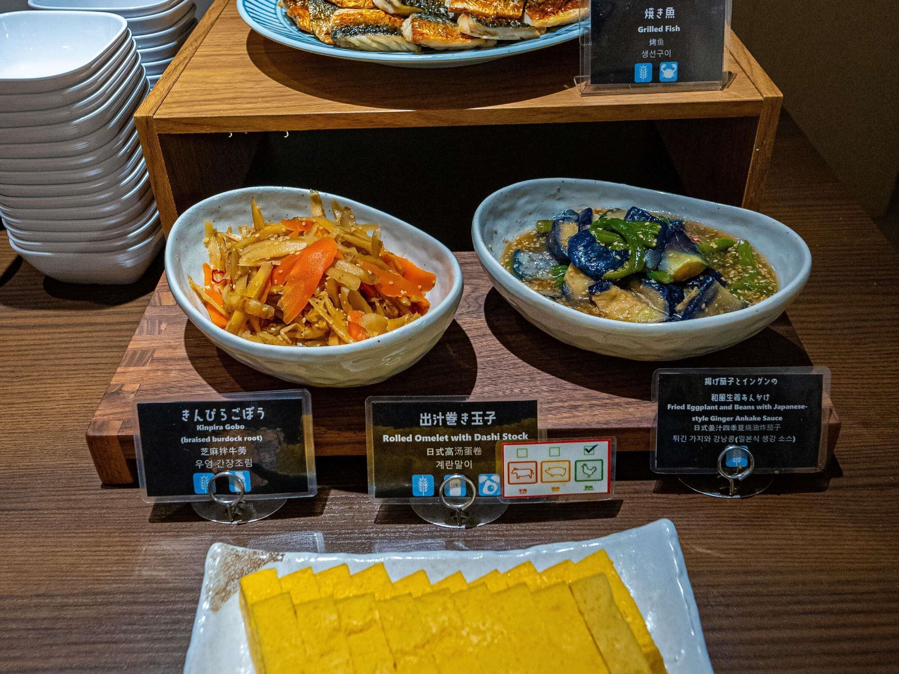 A buffet with various dishes, including a bowl of mixed vegetables, a bowl of fish, and a plate of yellow food.