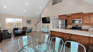 A spacious living area with a glass dining table and chairs, a kitchen with wooden cabinets and modern appliances, and a large TV mounted on the wall. - Prime Beach/Lagoon Location!  Walk to beach and restaurants! (Gulf Shores)