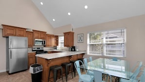 A modern kitchen with wooden cabinets, stainless steel appliances, a glass island, and a dining area with light blue chairs. - Prime Beach/Lagoon Location!  Walk to beach and restaurants! (Gulf Shores)