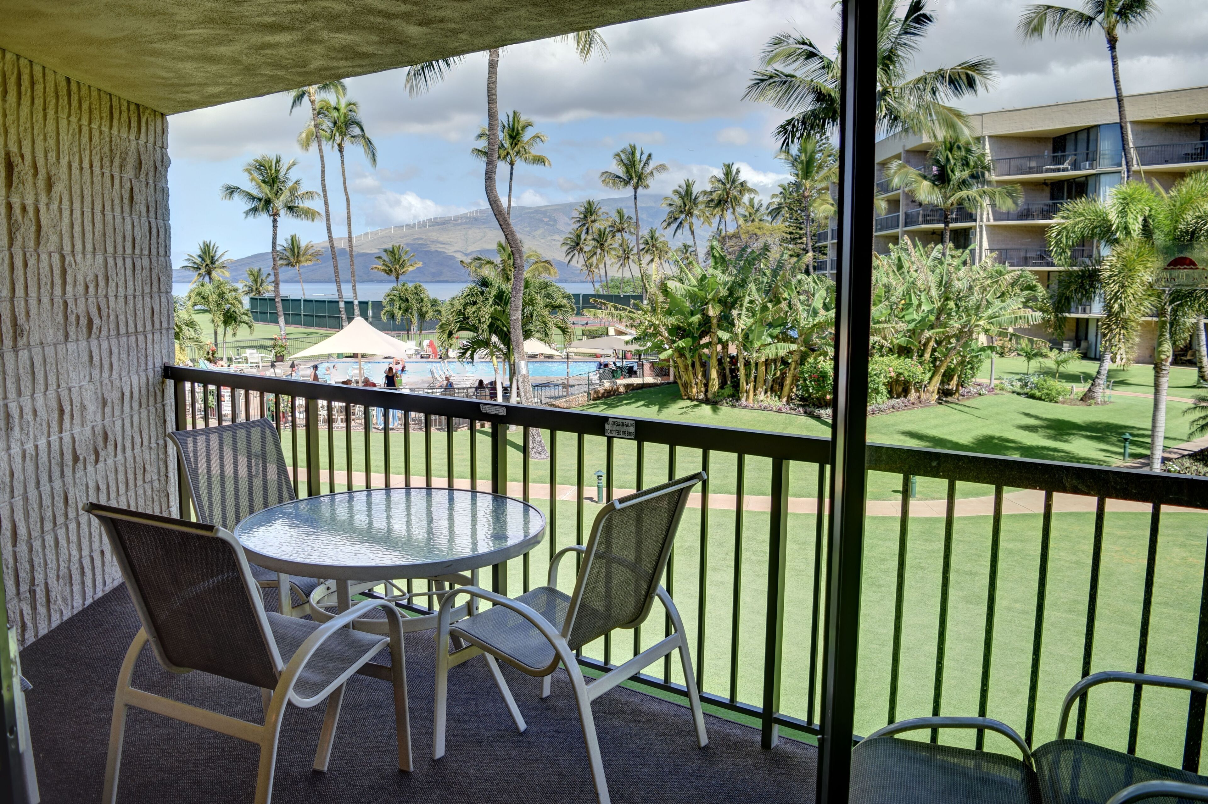 A balcony with a table and chairs, overlooking a pool area with palm trees and a building in the background.