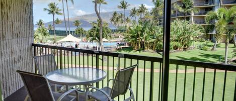 A balcony with a table and chairs, overlooking a pool area with palm trees and a building in the background.