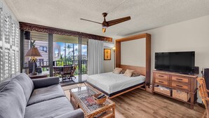 A modern hotel room with a balcony, a sofa, a bed, a TV, and a ceiling fan. - Beautiful garden view condo with tropical decor (Kihei)