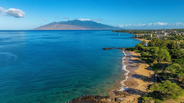 A coastal view with clear blue waters, a sandy beach, and a mountain in the background. - Beautiful garden view condo with tropical decor (Kihei)