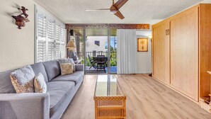 A modern living room with a grey sofa, a wicker coffee table, wooden flooring, a ceiling fan, and a sliding glass door leading to an outdoor patio. - Beautiful garden view condo with tropical decor (Kihei)