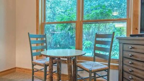 A dining area with a round table and two chairs, a wooden cabinet, and a large window with a view of greenery.