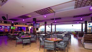 A spacious indoor dining area with multiple tables and chairs, a bar area with a television screen, and large windows offering a view of the outdoors. - Gold Crest Hotel - Mwanza (Mwanza)