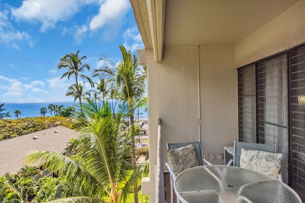 A balcony with a glass table and chairs, overlooking a beach with palm trees and a clear sky.