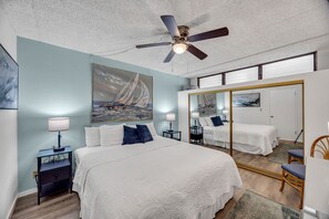 A bedroom with a large bed, a ceiling fan, a painting of a sailboat, a mirror, and a nightstand with a lamp.