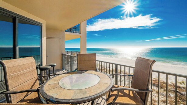 A balcony with a glass table and chairs, overlooking a beach and ocean.