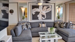 A modern living room with a grey sectional sofa, a glass coffee table, and a large abstract painting. - Oceanfront Condo at the Mana Kai Maui, Luxurious, Designer Remodeled (Kihei)