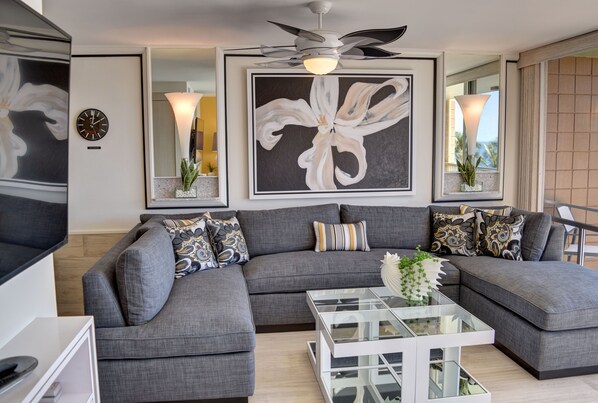 A modern living room with a grey sectional sofa, a glass coffee table, and a large abstract painting. - Oceanfront Condo at the Mana Kai Maui, Luxurious, Designer Remodeled (Kihei)