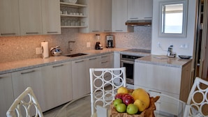 A modern kitchen with light wood cabinets, granite countertops, and a dining area with white chairs. - Oceanfront Condo at the Mana Kai Maui, Luxurious, Designer Remodeled (Kihei)