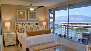 A bedroom with a bed, bedside tables, a bench, and a large window with a view of the sea and mountains. - Oceanfront Condo at the Mana Kai Maui, Luxurious, Designer Remodeled (Kihei)