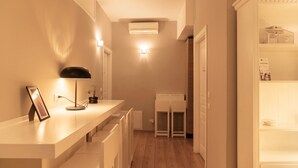 Un couloir étroit avec un comptoir blanc, un tableau encadré, une lampe sur pied et une petite plante.