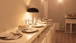 Une longue table à manger, dressée avec des assiettes, des couverts et des verres, entourée de chaises blanches, dans une pièce dotée d’un plancher en bois et d’une porte blanche.