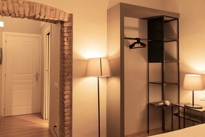 Habitación de hotel con suelo de madera, una puerta blanca, un arco de ladrillo, una estantería metálica negra y dos lámparas.