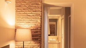 Un couloir avec un mur en briques et une lampe. Une porte donne accès à une salle de bain équipée d’un lavabo et d’un miroir.